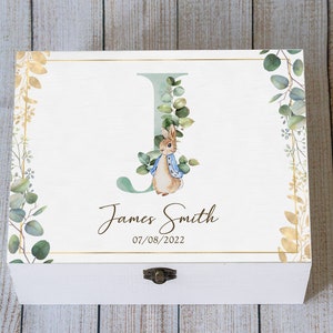 Op de afbeelding: Witte houten herinneringskist met een gouden rand en een aquarel illustratie van Peter Rabbit met de letter "J". De kist is gepersonaliseerd met de naam "James Smith" en de datum "07/08/2022". Decoratieve bloemaccenten omlijsten het ontwerp.