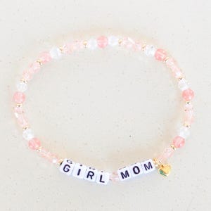 Può includere: Un braccialetto di perline rosa e trasparenti. Il braccialetto ha perline di lettere bianche che compongono la scritta "GIRL MOM" e un piccolo ciondolo a forma di cuore dorato. Le perline sono infilate su un cordino elastico trasparente.
