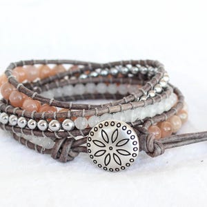 May include: Wrap-around bracelet with peach and white beads, and silver beads. The bracelet is secured with a grey leather strap and a silver button clasp with a floral design. The bracelet is made of multiple strands.