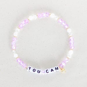 Può includere: Un braccialetto di perline rosa e bianche con un ciondolo dorato e le parole "YOU CAN" scritte in perline bianche.