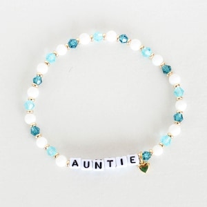 Può includere: Braccialetto di perline bianche e blu con la parola "AUNTIE" scritta in lettere bianche e un ciondolo a forma di cuore dorato.