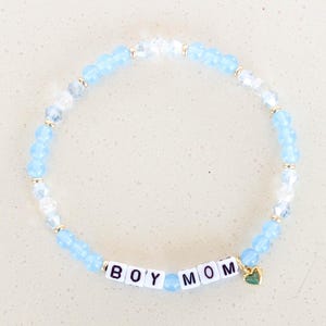 Può includere: Un braccialetto di perline blu chiaro con accenti dorati e un ciondolo. Il braccialetto ha le parole "BOY MOM" scritte in lettere bianche.