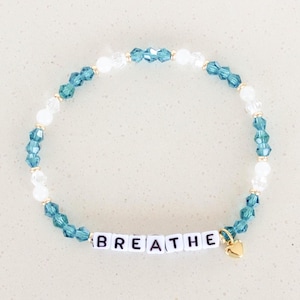 May include: A blue and white crystal beaded bracelet with a gold charm and the word "BREATHE" spelled out in white letters.