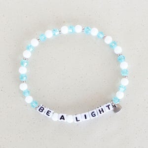 Può includere: Un braccialetto di perline bianche e azzurre. Il braccialetto presenta le parole "BE A LIGHT" scritte con perline a lettera bianche. Un piccolo ciondolo a forma di cuore argentato è attaccato.