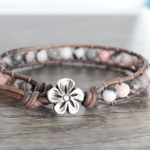 May include: Wrap bracelet with a silver flower clasp. The bracelet is made of brown leather and features grey, white, and pink beads. A fashionable accessory for any outfit.