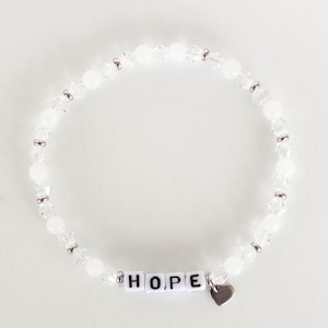May include: White beaded bracelet with the word "HOPE" spelled out in white letter beads. A small silver heart charm hangs from the bracelet. The bracelet is made of clear and white beads.