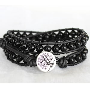 May include: A multi-wrap bracelet featuring two rows of polished black spherical beads strung on black leather cord. The bracelet includes a circular silver-toned button closure displaying a detailed purple line-art illustration of a tree.