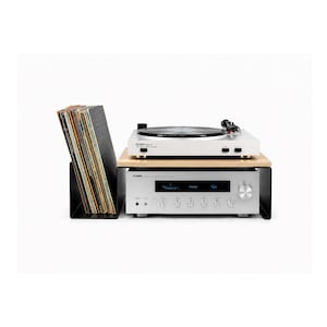 Puede incluir: Un tocadiscos blanco y un receptor estéreo plateado están apilados sobre una estantería de madera. A la izquierda hay un soporte negro para discos de vinilo, lleno de discos. La configuración está sobre un fondo blanco.