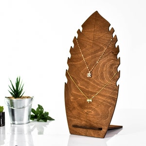 May include: A wooden leaf-shaped jewellery stand with two gold necklaces hanging from it. The stand has a brown wood finish and a small base.