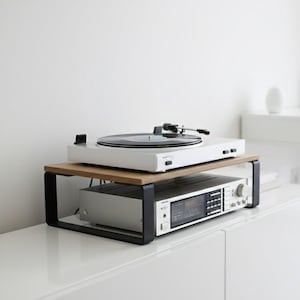 Könnte beinhalten: Ein weißer Plattenspieler steht auf einem Holzregal mit schwarzen Metallstützen, über einem Stereo-Receiver. Das Setup befindet sich auf einem weißen Schrank in einem minimalistischen, modernen Wohnraum.
