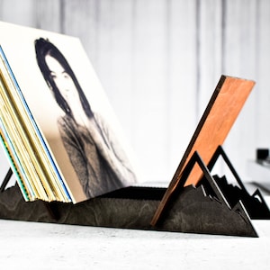 May include: A black wooden record stand with a mountain design. The stand holds a stack of vinyl records with a person's face on the cover.