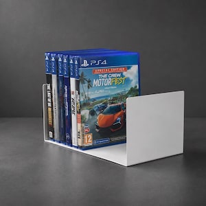 Może przedstawiać: Biały metalowy stojak w kształcie litery U mieszczący rząd siedmiu pudełek z grami wideo na PlayStation 4. Na przednim pudełku widnieje tytuł The Crew Motorfest Special Edition z pomarańczowym samochodem sportowym i niebieskim logo PS4.