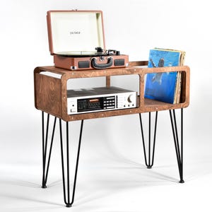 Può includere: Un supporto in legno per giradischi con gambe a forcina nere. Il supporto ha un ripiano integrato con un ricevitore stereo e una sezione per dischi in vinile. Un giradischi portatile marrone è in cima.