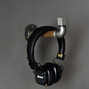 May include: A black headphone with a gold logo hanging on a silver pipe hook attached to a wooden base. The hook is mounted on a gray wall.