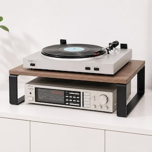 Op de afbeelding: Een zilveren platenspeler met een zwarte plaat staat op een houten plank met zwarte metalen poten. Daaronder is een zilveren stereoreceiver zichtbaar. De opstelling staat op een wit oppervlak.