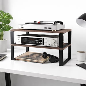 May include: A two-tier audio equipment stand featuring dark brown wood-grain shelves supported by matte black metal frames. The unit holds a silver turntable on the top shelf and a silver stereo receiver on the bottom shelf, with a vinyl record underneath.