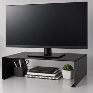 Op de afbeelding: Een matzwarte metalen monitorstandaard met een gebogen ontwerp, waarop een platte computermonitor staat. Onder de standaard bevinden zich een zwarte vierkante pennenhouder met pennen, een stapel van drie notitieboekjes en een kleine vetplant in een pot.