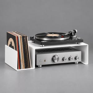 May include: A white acrylic storage stand with a side compartment for vinyl records. A black turntable sits on the top surface, while a silver audio amplifier with four circular control knobs and a headphone jack is positioned underneath the stand.