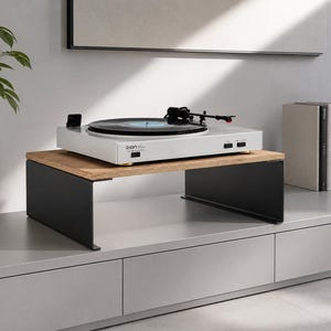Puede incluir: Un tocadiscos blanco con un brazo negro se encuentra sobre un soporte de madera y negro. El soporte está colocado sobre un mueble gris. También se ven una planta en maceta, libros y una lámpara gris.