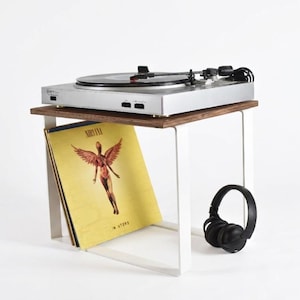 May include: A silver turntable on a small wooden table with white metal legs. A yellow album cover with a winged figure and the word "NIRVANA" is propped against the table's side. Black headphones are also present.