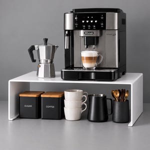 Może przedstawiać: Ekspres do kawy De'Longhi Magnifica S stoi na białej półce wraz ze srebrną kawiarką i szklanką latte. Pod półką znajdują się dwa czarne pojemniki z napisami SUGAR i COFFEE, trzy beżowe kubki, czarny dzbanek oraz złote łyżeczki.