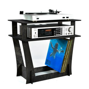 Puede incluir: Un soporte de madera negro para tocadiscos con dos estantes. El estante superior tiene una tornamesa y un receptor estéreo. El estante inferior tiene una pila de discos de vinilo con un álbum de Nirvana visible.