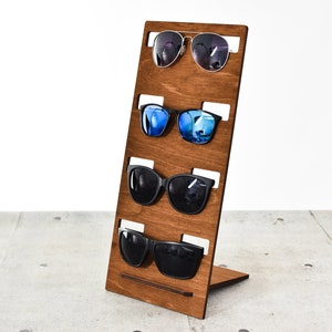 Puede incluir: Un soporte de madera con cinco ranuras para exhibir gafas de sol. El soporte es marrón y tiene un acabado de madera natural. Las gafas de sol son de diferentes estilos y colores.