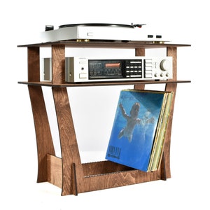 Puede incluir: Un soporte de tocadiscos de madera con dos estantes. El estante superior tiene una plataforma giratoria y un receptor estéreo. El estante inferior tiene una pila de discos de vinilo, incluyendo uno con una cubierta azul que muestra a un niño nadando bajo el agua. El texto "NIRVANA" e "IN UTERO" es visible en la portada del disco.