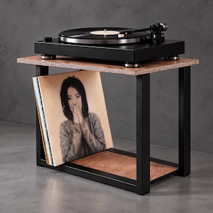 Può includere: Un giradischi nero è posizionato su un supporto rettangolare in legno con una struttura in metallo nero. Il supporto è dotato di un ripiano inferiore che ospita una collezione di dischi in vinile, tra cui uno con il ritratto di una persona.