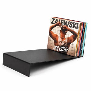 Może przedstawiać: Matowy czarny metalowy stojak na płyty winylowe z płaską podstawą i pionowym oparciem. Kolekcja płyt winylowych ustawiona jest pionowo przy oparciu, a przednia okładka albumu zawiera napis ZALEWSKI ZGŁOWY oraz wizerunek osoby.