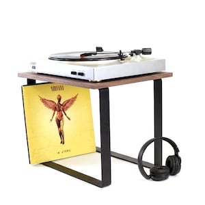 Puede incluir: Un tocadiscos blanco sobre una pequeña mesa de madera con patas de metal negro. Un álbum de vinilo con una figura alada se muestra en el estante de abajo. Los auriculares negros descansan a un lado.