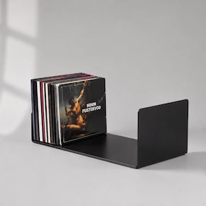 Könnte beinhalten: Ein U-förmiger Schallplattenständer aus schwarzem Metall, der eine Sammlung von Schallplatten hält. Das vorderste Plattencover zeigt ein dunkles klassisches Gemälde und den weißen Text MININ PUSTERVOD. Der Ständer hat eine flache Basis und zwei Seiten.
