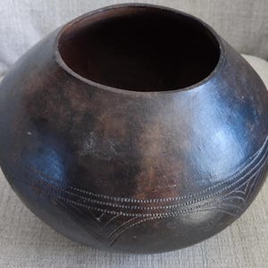 May include: A large, dark brown, round ceramic pot with a wide opening and a decorative pattern of incised lines around the rim and bottom.