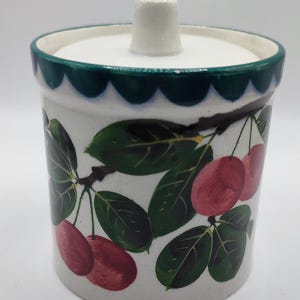 May include: A ceramic jar with a lid, decorated with a cherry and leaf design. The jar has a white base, a green scalloped rim, and a white lid with a small knob. The design features red cherries and green leaves.