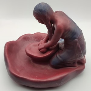 Op de afbeelding: Een kastanjebruin en blauw getint beeldje van een persoon die op de knieën zit en met klei werkt op een ronde pottenbakkersschijf. De figuur heeft lang haar en buigt zich over een kleine, ronde kom op een brede, getextureerde ondergrond.