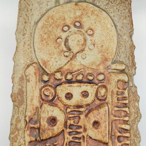 Puede incluir: Un colgante de pared rectangular de cerámica texturizada, con una paleta de colores beige y marrón. El diseño presenta un gran elemento circular en la parte superior con pequeñas hendiduras circulares y formas geométricas debajo. Los bordes tienen una textura rugosa.