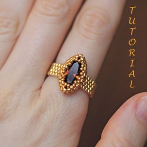 K&ouml;nnte beinhalten: Ein goldfarbener Perlenring mit einem maronenfarbenen, markisenf&ouml;rmigen Edelstein. Der Ring befindet sich an einem Finger.