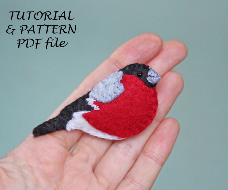 May include: A felt bird brooch with a red body, grey head, and black wings. The bird is sitting on a hand.