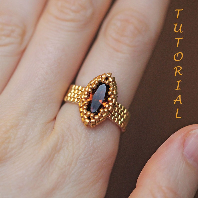 K&ouml;nnte beinhalten: Ein goldfarbener Perlenring mit einem maronenfarbenen, markisenf&ouml;rmigen Edelstein. Der Ring befindet sich an einem Finger.