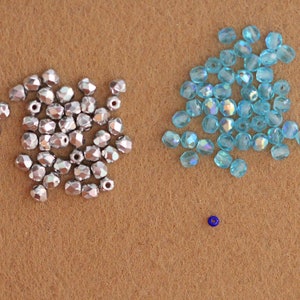 May include: A close-up of two piles of beads on a brown surface. One pile is silver and the other is light blue. There is a single blue bead in the center of the image.
