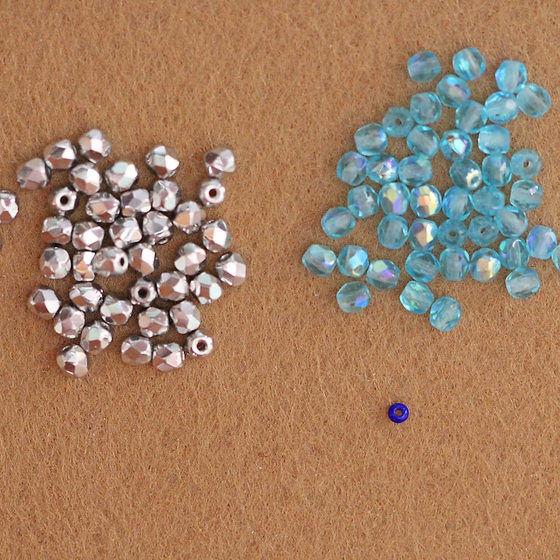May include: A close-up of two piles of beads on a brown surface. One pile is silver and the other is light blue. There is a single blue bead in the center of the image.