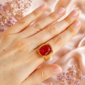 May include: A ring featuring a large, rectangular, faceted red gemstone set within a gold-toned beaded frame. The band of the ring is constructed from small, uniform gold-colored beads arranged in a woven, grid-like pattern around the finger.
