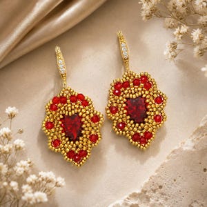 May include: A pair of drop earrings featuring a central teardrop-shaped red crystal surrounded by intricate gold and red beadwork. The earrings hang from gold hooks accented with small clear crystals, creating a detailed floral-like design.