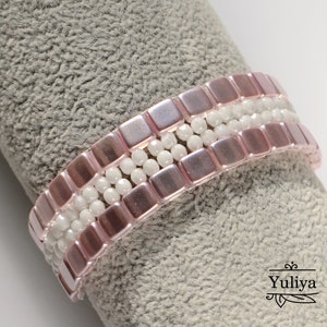 May include: A pink and white beaded bracelet with square and round beads. The bracelet is on a gray fabric background.