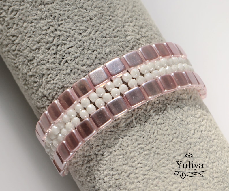 May include: A pink and white beaded bracelet with square and round beads. The bracelet is on a gray fabric background.