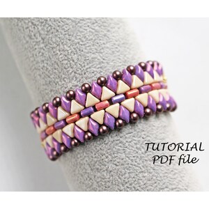 Op de afbeelding: Een paarse, crème en roze kralenarmband met een geometrisch patroon. De armband is gemaakt met kleine, driehoekige kralen en ligt op een grijs oppervlak. De tekst "TUTORIAL PDF file" is zichtbaar in de afbeelding.