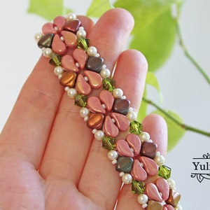 May include: A beaded bracelet with pink, brown, and green beads in a floral pattern. The bracelet is on a hand, with green foliage in the background.