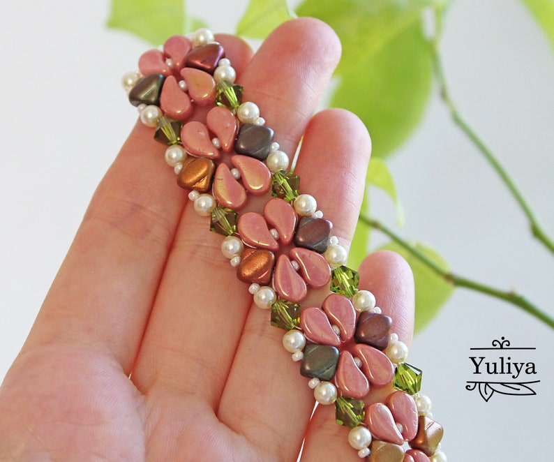 May include: A beaded bracelet with pink, brown, and green beads in a floral pattern. The bracelet is on a hand, with green foliage in the background.