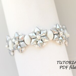 Puede incluir: Un brazalete de cuentas plateadas y blancas con un dise&ntilde;o de copo de nieve. El brazalete est&aacute; hecho con cuentas peque&ntilde;as y redondas y cuentas m&aacute;s grandes y facetadas. El brazalete est&aacute; sobre un fondo blanco. TUTORIAL PDF file.