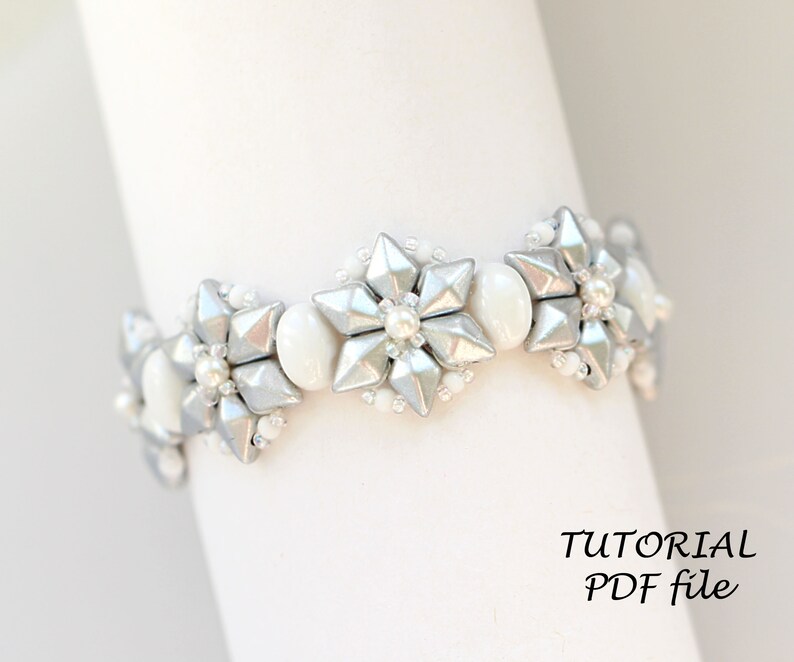 Puede incluir: Un brazalete de cuentas plateadas y blancas con un dise&ntilde;o de copo de nieve. El brazalete est&aacute; hecho con cuentas peque&ntilde;as y redondas y cuentas m&aacute;s grandes y facetadas. El brazalete est&aacute; sobre un fondo blanco. TUTORIAL PDF file.